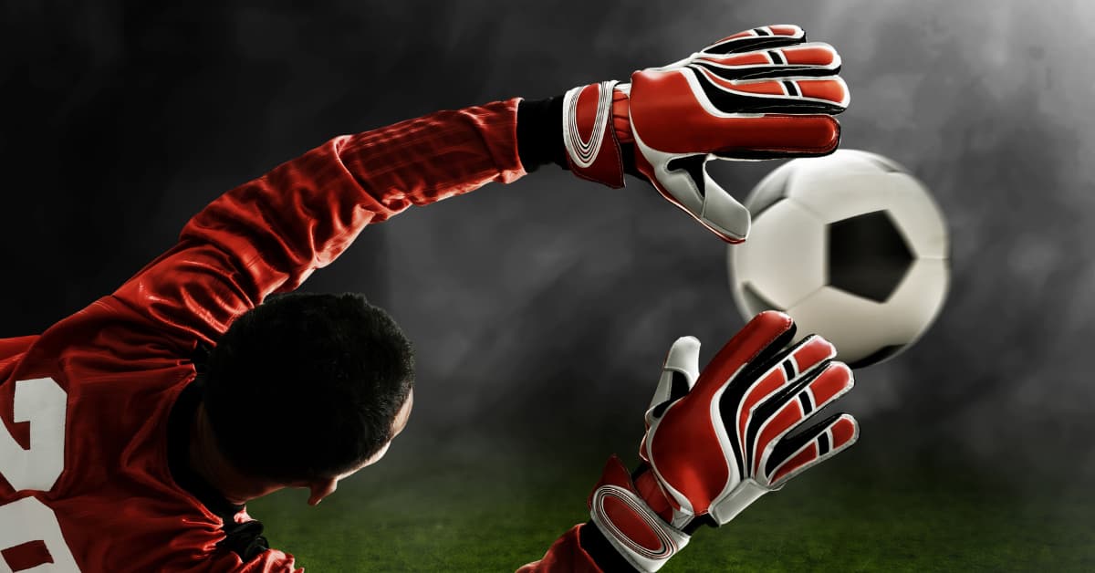 Portero con guantes de portero rojos deteniendo un balón en pleno vuelo durante un partido de fútbol.
