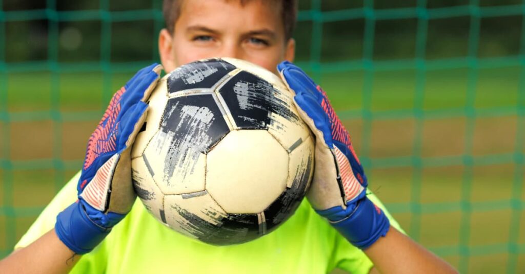 Niño portero sujetando un balón con sus primeros guantes de portero frente a la portería.